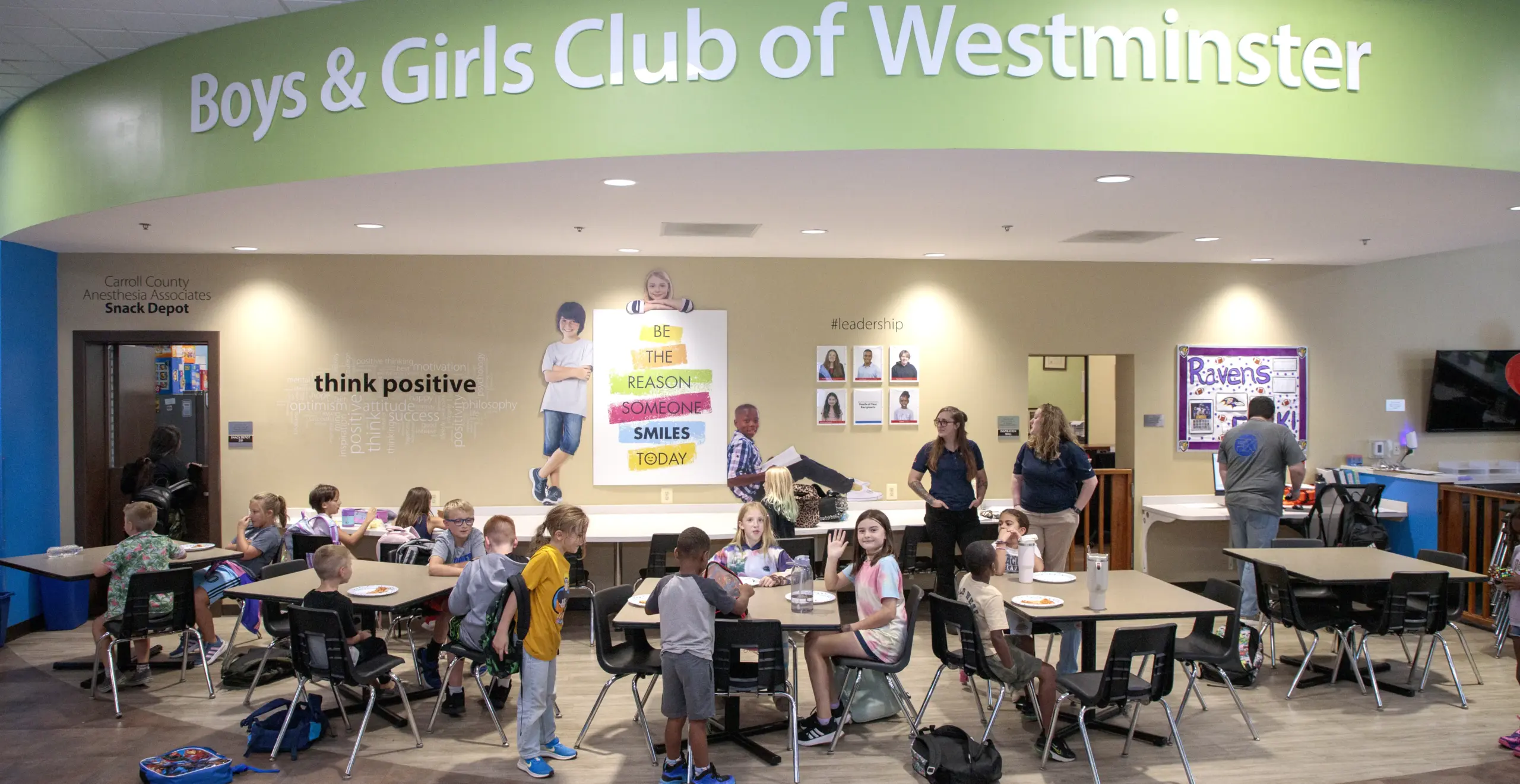 Cafeteria scene at Boys & Girls Club of Westminster with groups of children seated at tables eating, and a few adults standing near the beige wall with posters and a large green curved sign overhead reading ’Boys & Girls Club of Westminster.’
