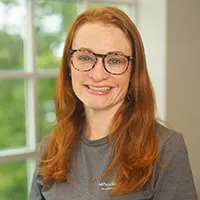 Portrait of a smiling woman with long red hair and glasses, wearing a gray shirt, indoors near a window at the backlight.