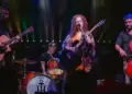 Live band on stage: a female guitarist/vocalist center stage with two guitarists and a cellist, drummer in back, colorful stage lights behind them