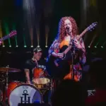 Ebb & Nova - Carroll News | Carroll Publications | Carroll County, MD Live band on stage: a female guitarist/vocalist center stage with two guitarists and a cellist, drummer in back, colorful stage lights behind them