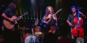 Live band on stage: a female guitarist/vocalist center stage with two guitarists and a cellist, drummer in back, colorful stage lights behind them