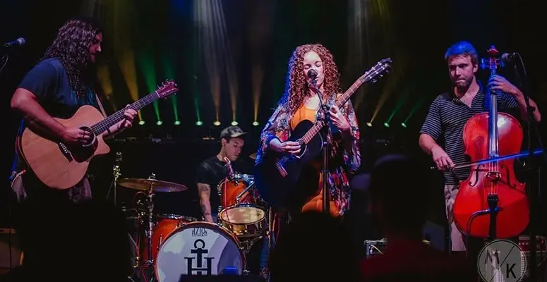 Live band on stage: a female guitarist/vocalist center stage with two guitarists and a cellist, drummer in back, colorful stage lights behind them