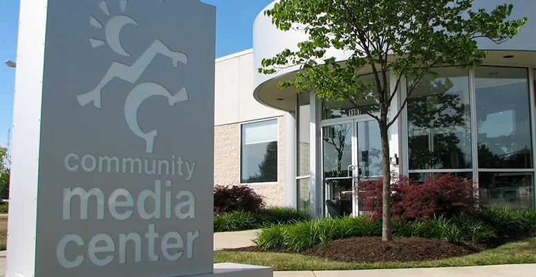 Front view of a community media center with a tall sign reading 'community media center' and a landscaped entrance with glass doors and trees.