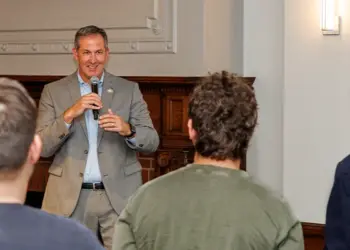 Man in a gray suit speaks into a microphone to a small audience in a conference room