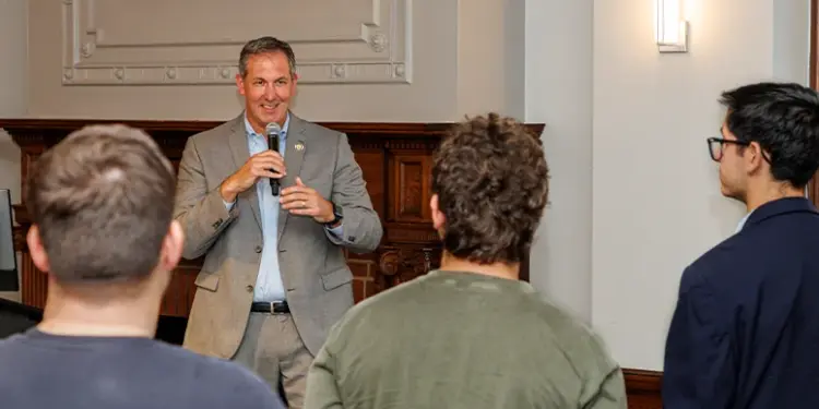 Man in a gray suit speaks into a microphone to a small audience in a conference room