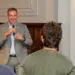 Man in a gray suit speaks into a microphone to a small audience in a conference room