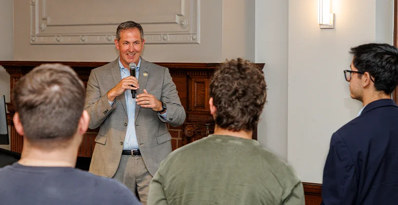Man in a gray suit speaks into a microphone to a small audience in a conference room