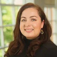 Portrait of a smiling woman with long brown hair, wearing a black turtleneck, indoors near a window lighted by greenery.