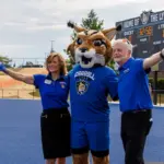 Mince, Lynx, Ball_FeaturedImage - Carroll News | Carroll Publications | Carroll County, MD Mascot in a blue uniform with two staff members on a blue track, arms raised with pom-poms; scoreboard in the background reads 'Home of the Lynx'.