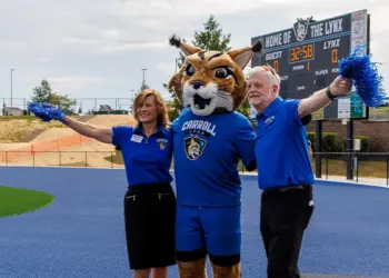 Mascot in a blue uniform with two staff members on a blue track, arms raised with pom-poms; scoreboard in the background reads 'Home of the Lynx'.