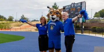 Mascot in a blue uniform with two staff members on a blue track, arms raised with pom-poms; scoreboard in the background reads 'Home of the Lynx'.