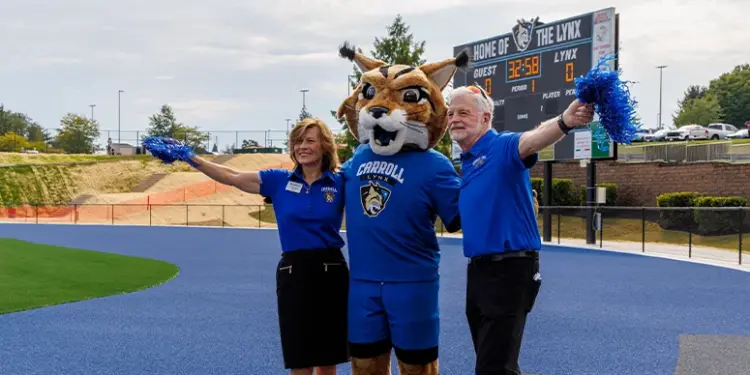 Mascot in a blue uniform with two staff members on a blue track, arms raised with pom-poms; scoreboard in the background reads 'Home of the Lynx'.