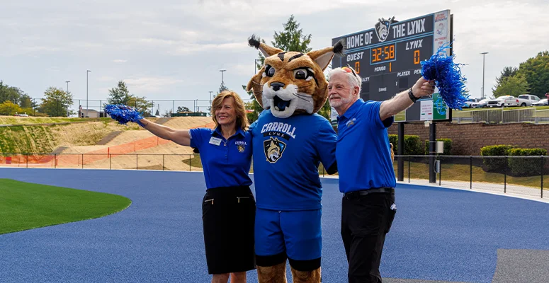 Mascot in a blue uniform with two staff members on a blue track, arms raised with pom-poms; scoreboard in the background reads 'Home of the Lynx'.