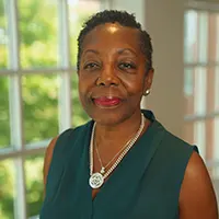 Portrait of a smiling woman wearing a teal sleeveless top and pearl necklace, indoors by a window.