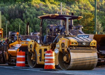 Row of yellow Caterpillar road rollers at a construction site, orange traffic barrels in the foreground, with a forested hillside in the background.