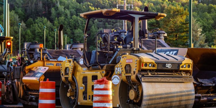 Row of yellow Caterpillar road rollers at a construction site, orange traffic barrels in the foreground, with a forested hillside in the background.