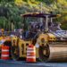 Row of yellow Caterpillar road rollers at a construction site, orange traffic barrels in the foreground, with a forested hillside in the background.