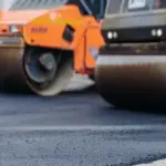 Two large road rollers compacting fresh asphalt on a construction site, with a worker in a reflective vest nearby.