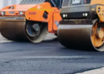 Two large road rollers compacting fresh asphalt on a construction site, with a worker in a reflective vest nearby.