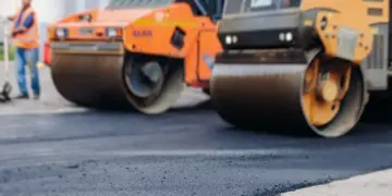 Two large road rollers compacting fresh asphalt on a construction site, with a worker in a reflective vest nearby.