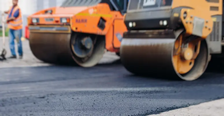 Two large road rollers compacting fresh asphalt on a construction site, with a worker in a reflective vest nearby.