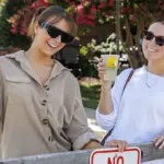 Kayla-Willing-Hannah-Curtright - Carroll News | Carroll Publications | Carroll County, MD Two women wearing sunglasses lean on a wooden fence, holding lemonades and smiling outdoors at a sunny event.",
