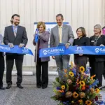 Ribbon Cutting feature image - Carroll News | Carroll Publications | Carroll County, MD Group of ten adults in business attire participate in a ribbon-cutting ceremony for Carroll Community College, with a blue ribbon and a colorful flower arrangement at the front.