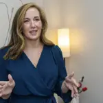 Stephanie Powers Q&A - Carroll News | Carroll Publications | Carroll County, MD Woman in a blue wrap dress speaks with open hands in a room with butterfly mural and a lamp behind her.
