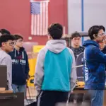 ccps-career-tech-image - Carroll News | Carroll Publications | Carroll County, MD Group of students in a machine shop standing around workbenches and equipment, discussing a project, with an American flag in the background.